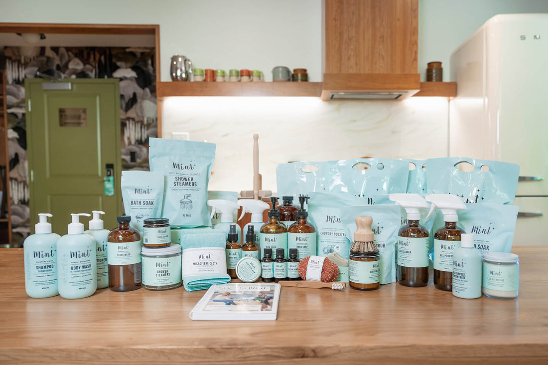 Mint Cleaning Products displayed on a wooden countertop, featuring eco-friendly body care, cleaning sprays, and laundry items in light mint packaging, with a kitchen backdrop.