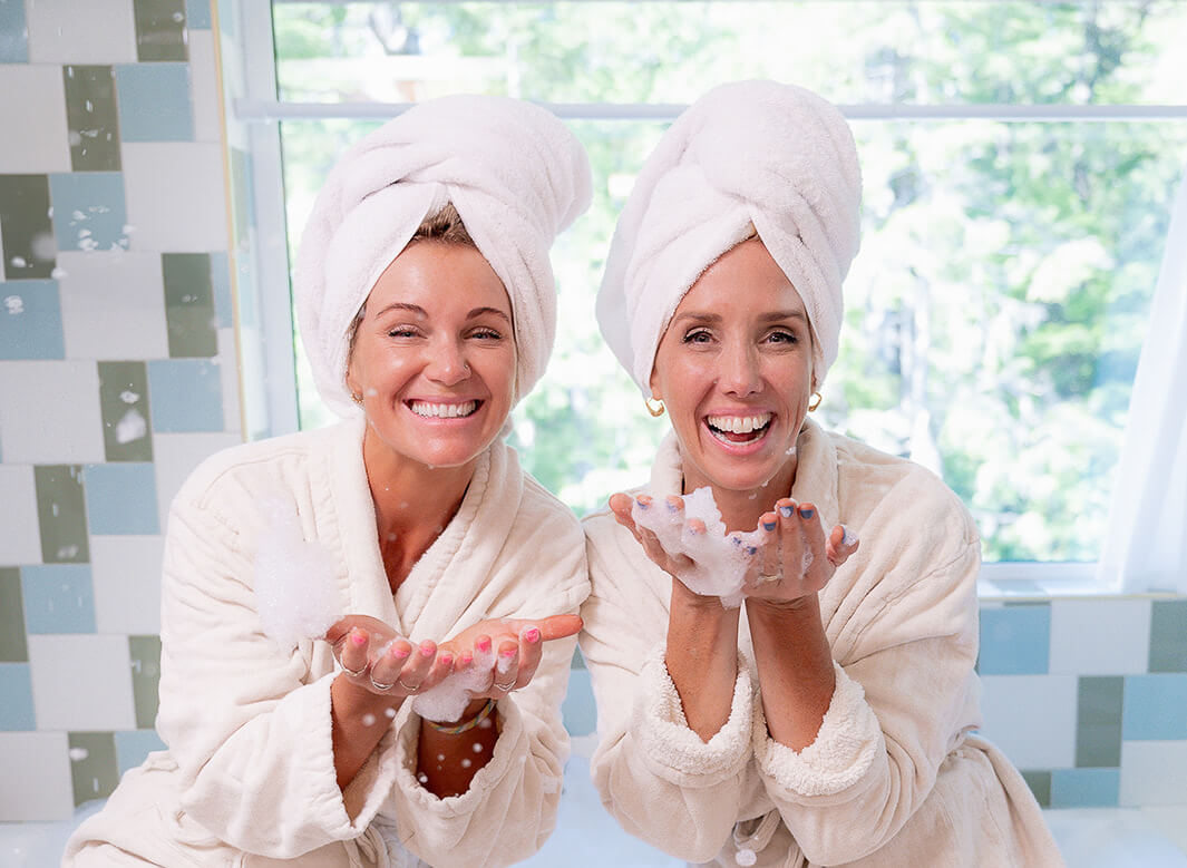 Monika and Robyn, co-founders of Mint Cleaning Products, smiling in bathrobes with Mint Body Care products displayed behind them in a bright, tiled bathroom.