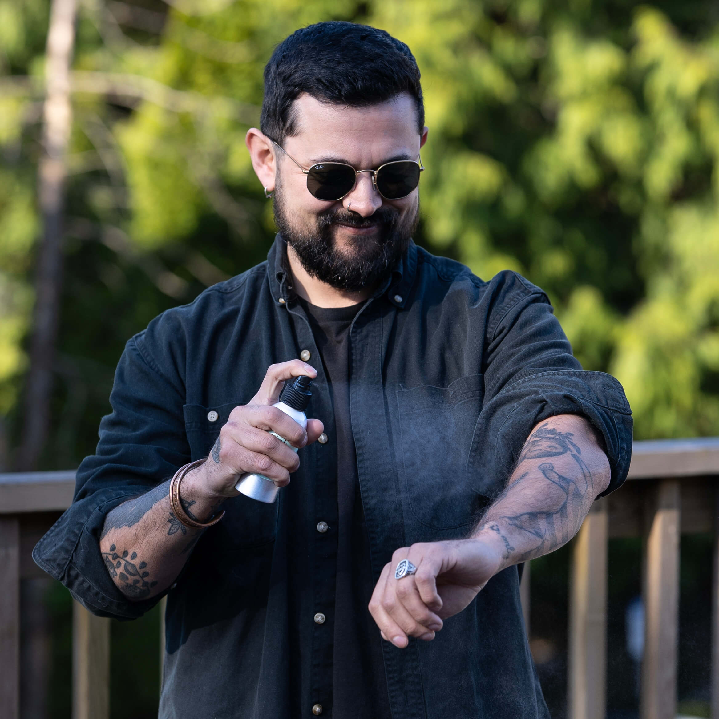 A man wearing sunglasses and a black shirt sprays Mint Outdoor Mist onto his arm while standing outdoors on a deck. The forest is visible in the background.